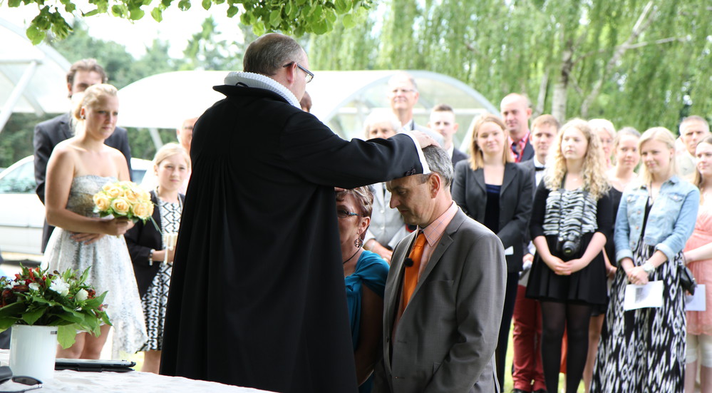 Præst velsigner et brudepar udenfor med hænderne på deres hoveder, mens gæsterne ser på