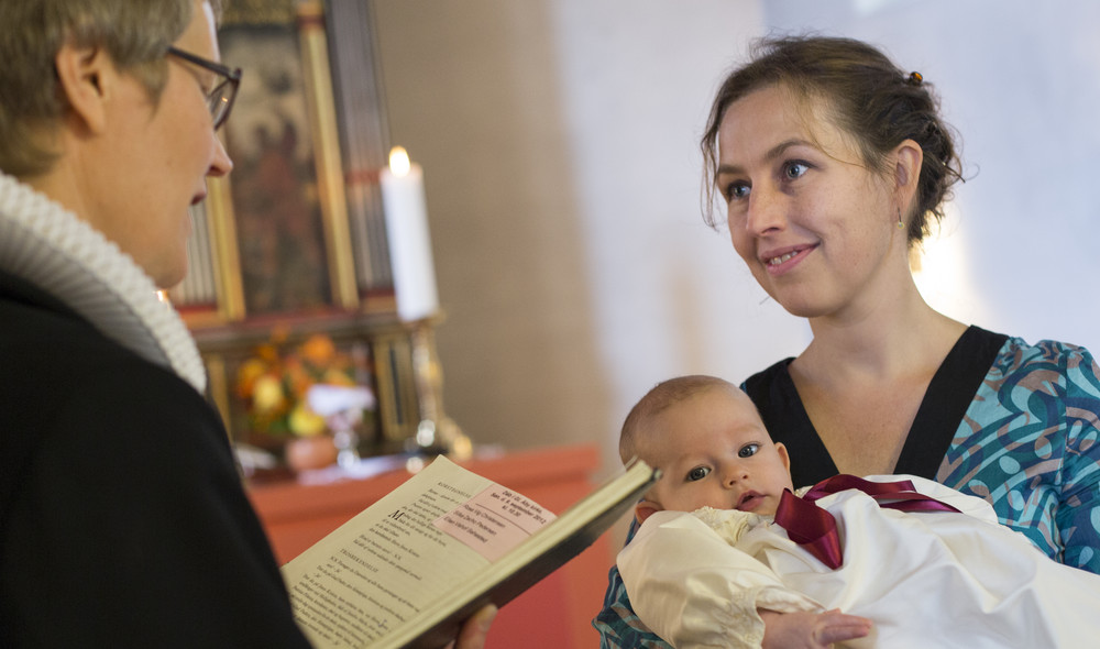 Gudmor med dåbsbarn i dåbskjole smiler til præst, der læser højt fra ritualbogen til barnedåb i folkekirken